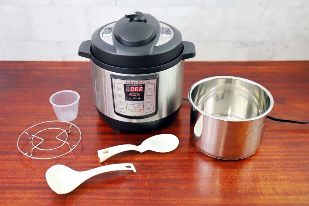 A multi-function electric pressure cooker with accessories, including a metal inner pot, plastic measuring cup, trivet, and two white serving spoons, on a wooden surface.