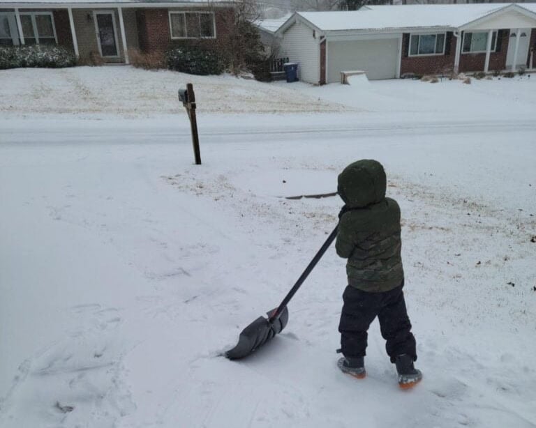 A child in winter clothing shovels snow from a driveway in a residential neighborhood under an overcast sky.