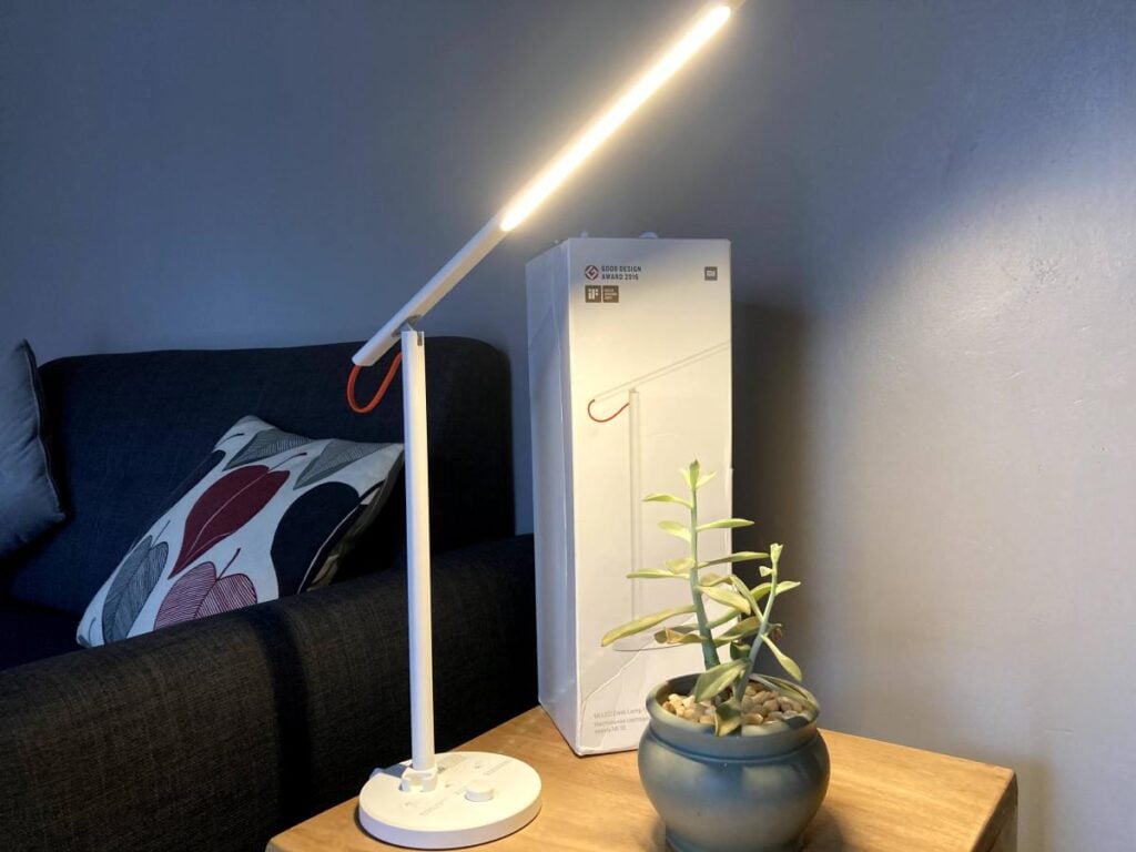 A lit desk lamp stands on a table beside a small potted plant and a white box. A couch with patterned pillows is in the background.