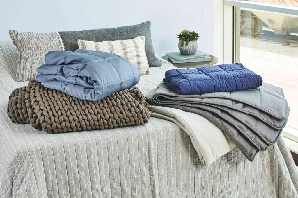 A bed with folded blankets and comforters in various colors and textures. A small potted plant sits on a side table by the window.