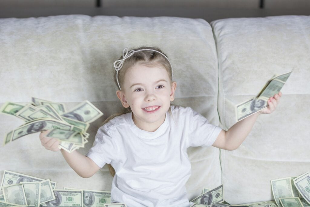 Young child sitting on a couch, smiling and holding multiple U.S. dollar bills, with more scattered around.