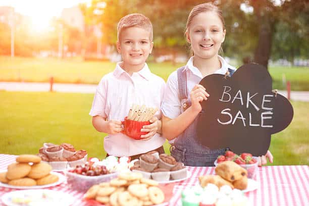 Two children stand at a bake sale table outdoors. One holds a sign reading 