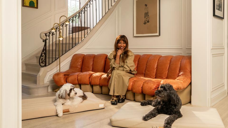  Brigette Romanek sitting on an orange couch in a grand entryway. There is a white dog and a black dog both lay on her designer dog beds. 