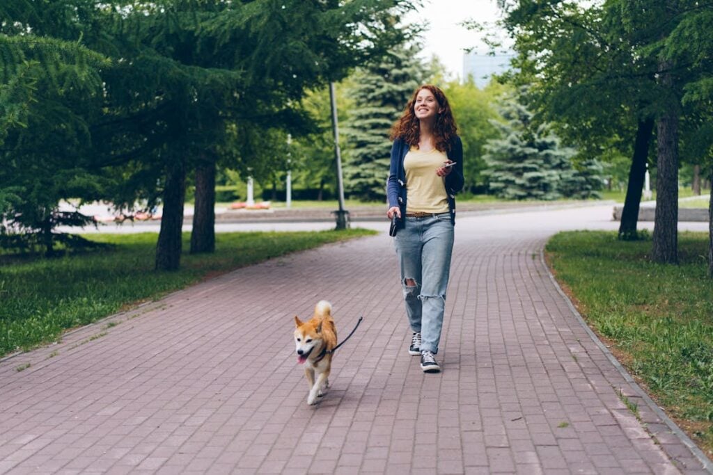 A woman walks a small dog on a leash along a park path surrounded by trees, with a joyful expression.