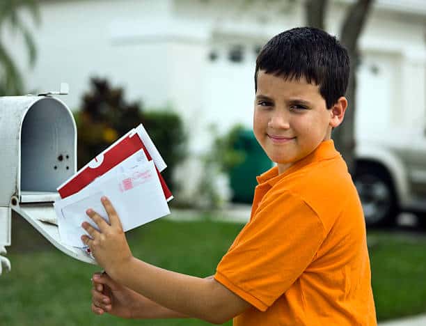 A boy in an orange shirt stands by an open white mailbox, holding several pieces of mail.