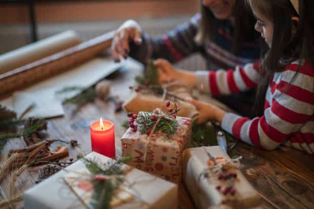 A person and a child wrap Christmas gifts at a wooden table with a lit red candle and natural decorations like pine branches and berries.