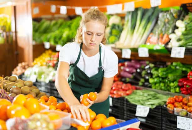 A woman in a green apron arranges oranges in a grocery store's produce section. Various fruits and vegetables are displayed in the background.