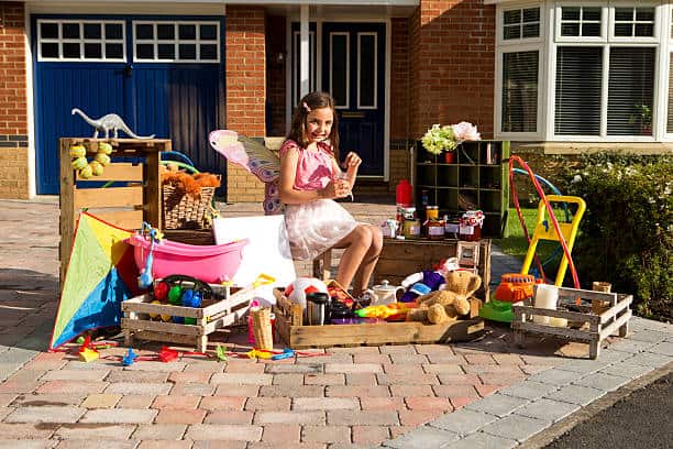 A girl in fairy wings sits on a crate surrounded by toys, jars, and a kite in a driveway sale setup.