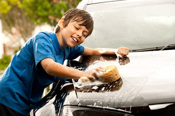 A boy in a blue shirt is smiling while washing a car with a sponge, surrounded by soap suds.