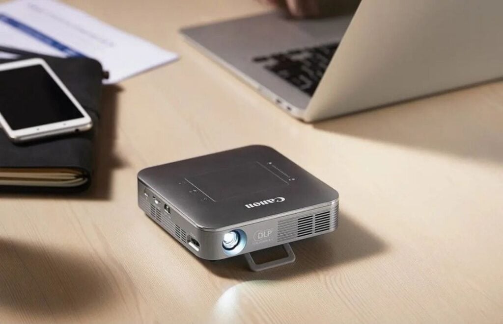 A Canon DLP projector sits on a wooden desk, next to a laptop and a smartphone.