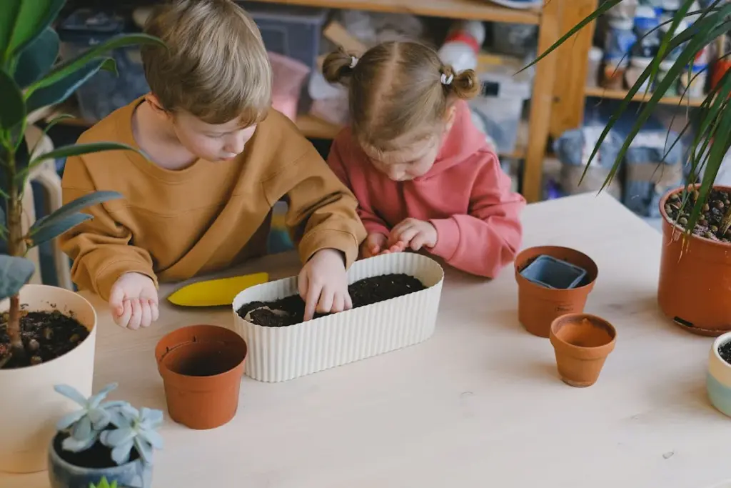 Two children planting seeds in a rectangular pot on a table with several other small pots around them.