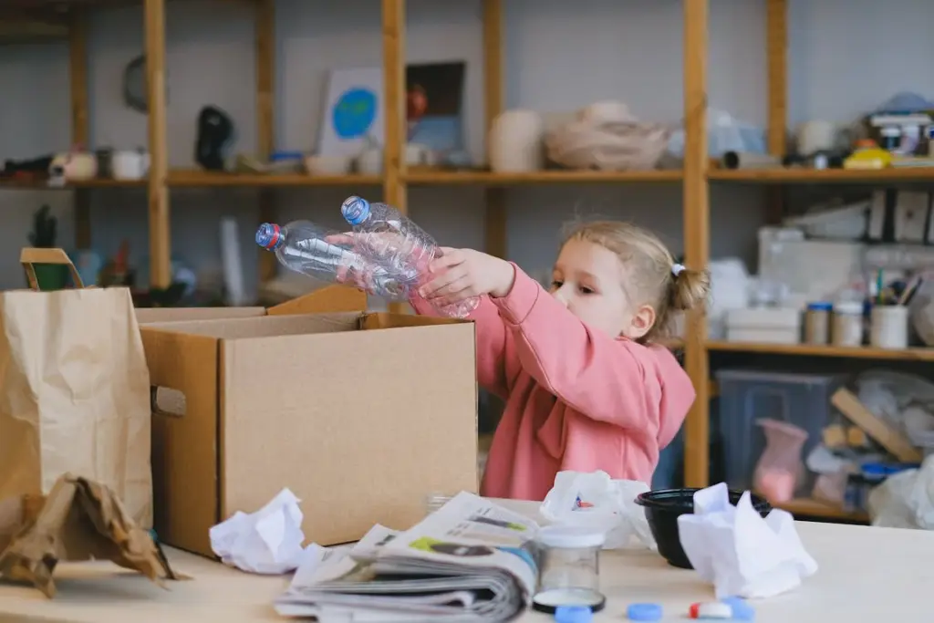 Child placing plastic bottles into a cardboard box surrounded by papers and recyclables in a room with shelves in the background.