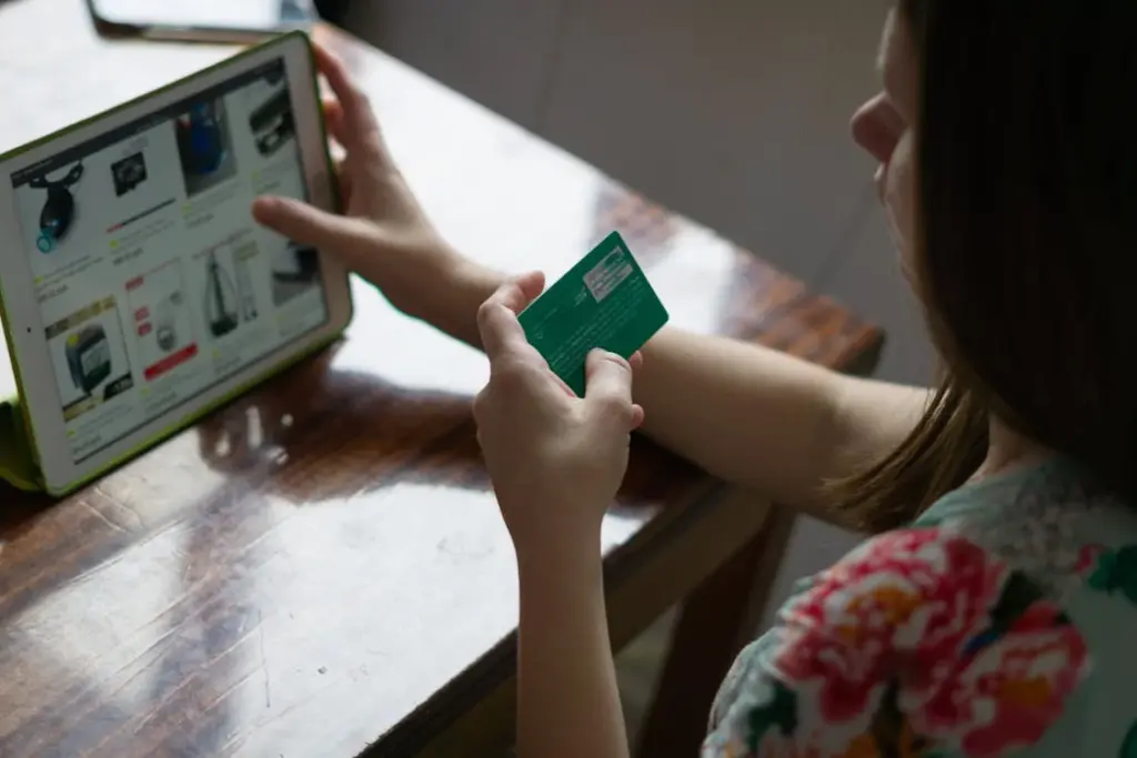 Person holding a credit card while shopping online on a tablet at a wooden table.