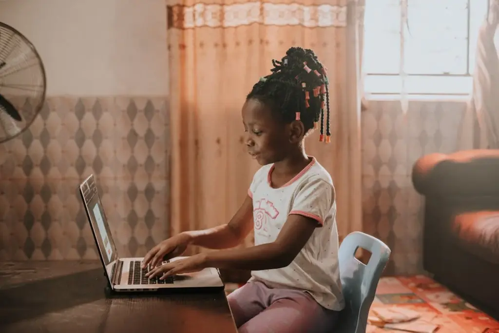 A young girl sits at a table using a laptop in a cozy room with soft lighting.