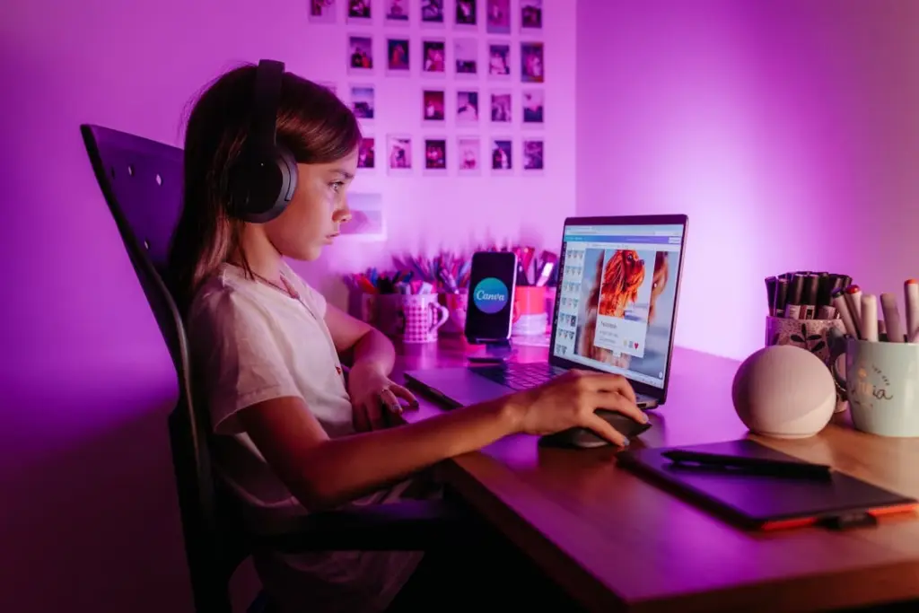 A girl wearing headphones works on a laptop at a desk, surrounded by stationery and a smart speaker, under pink lighting with photo wall in background.