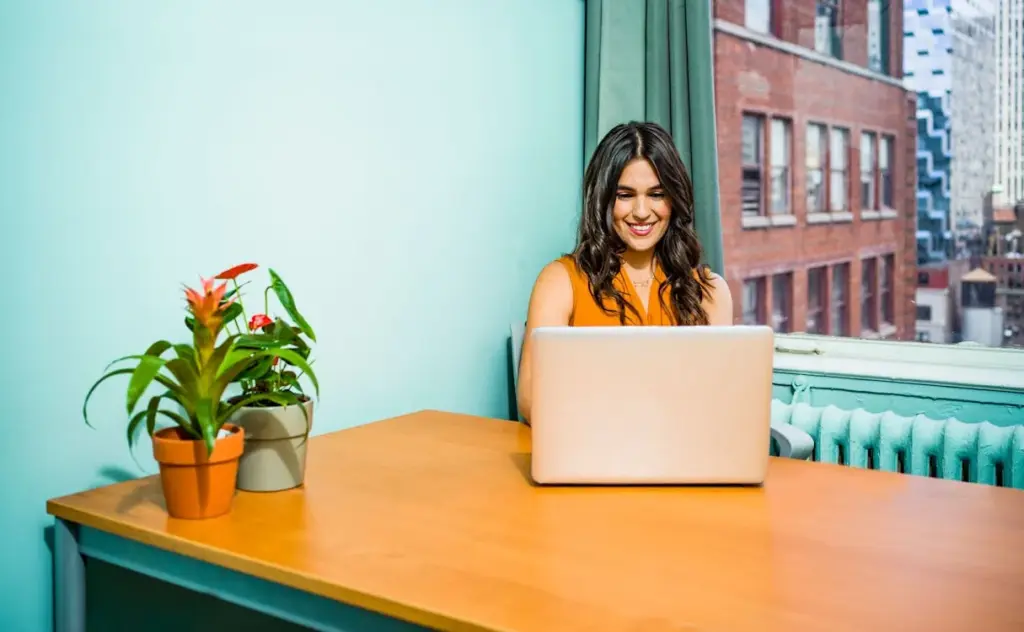 A woman in an orange top sits at a desk using a laptop, with potted plants beside her and a cityscape visible through the window.