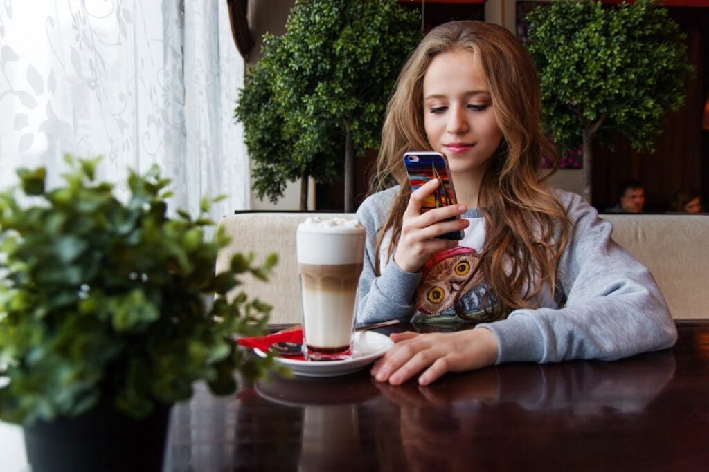 A young woman sits at a café table, looking at her smartphone. A latte and a small plant are on the table in front of her.