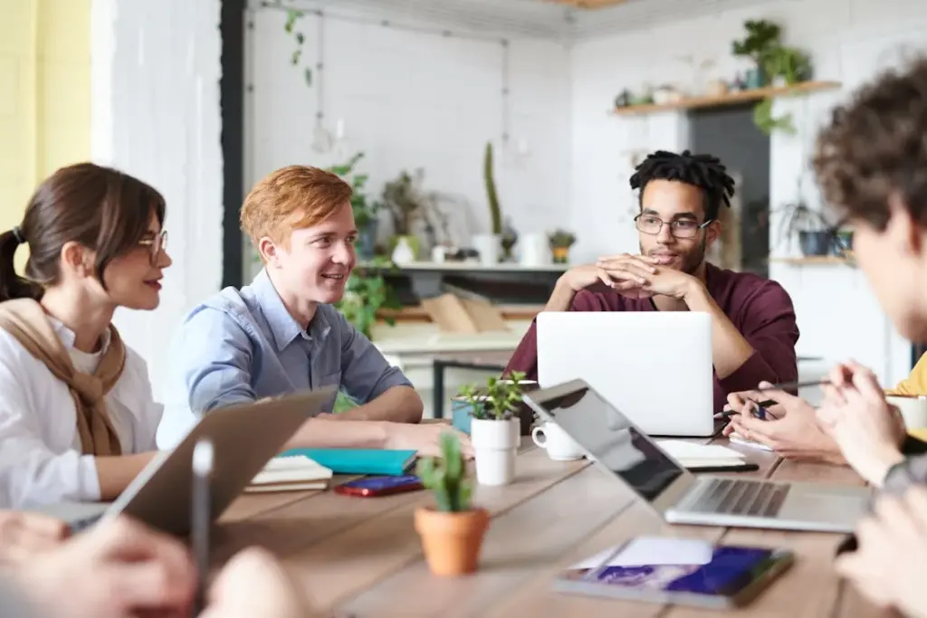 A group of people sitting around a table with laptops and notebooks, engaged in a meeting in a well-lit room with plants in the background.