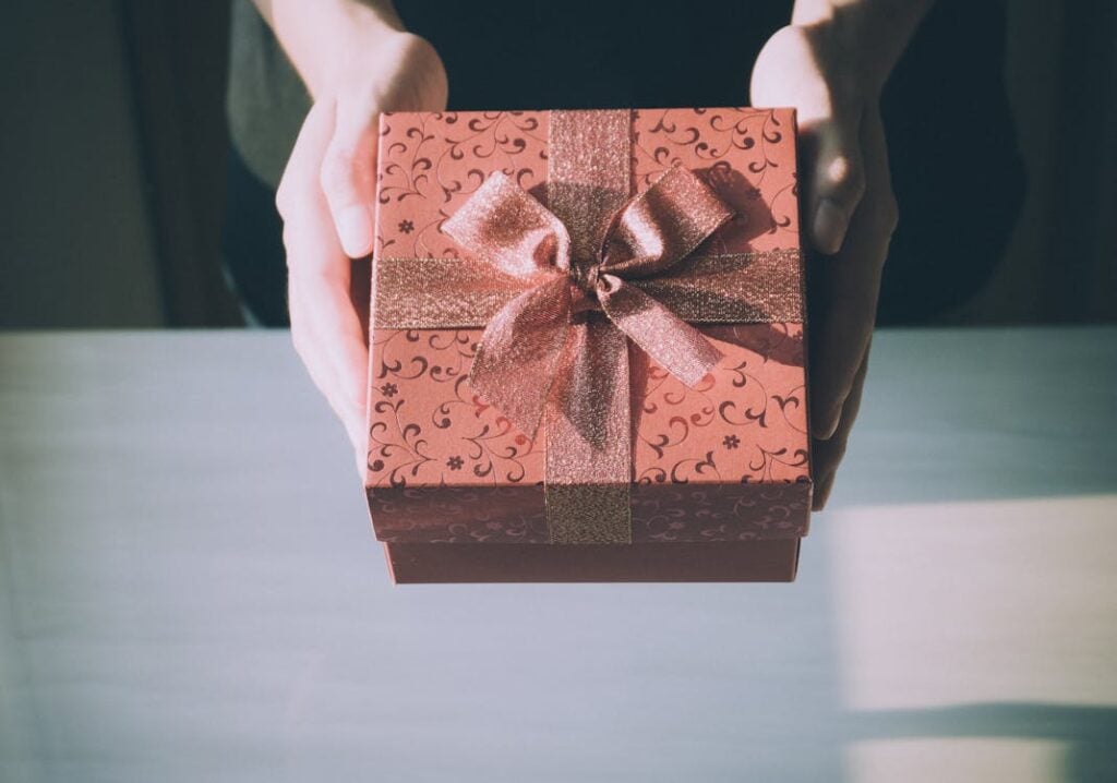 Hands holding a decorated gift box with a patterned design and a shiny ribbon bow on top.