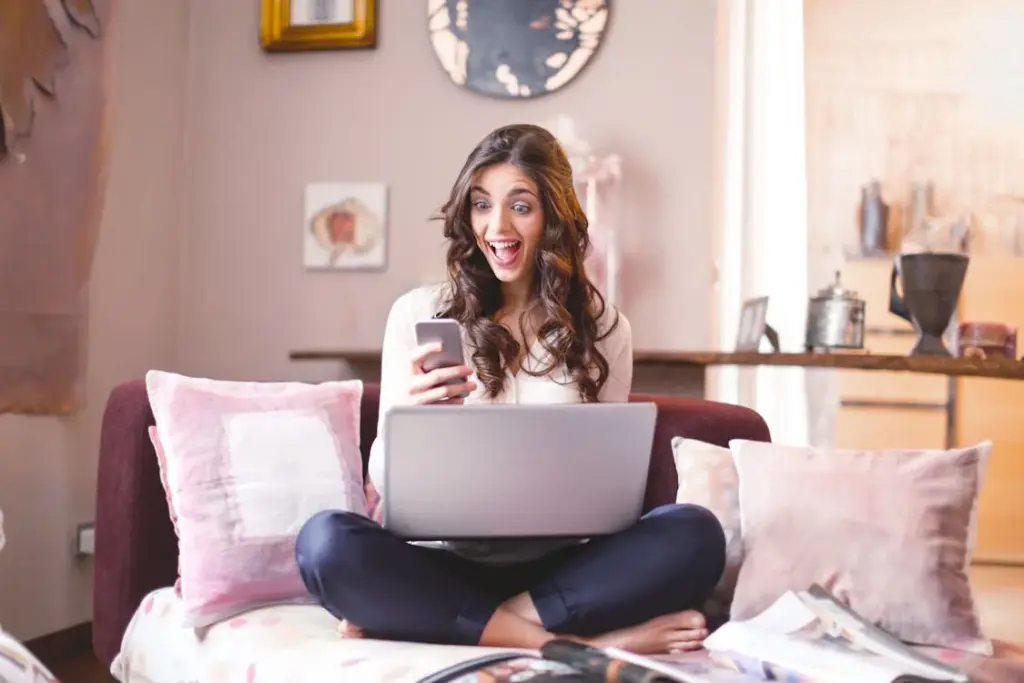 A woman sitting on a couch with a laptop on her lap looks at her phone screen with a surprised expression. She is surrounded by pillows and a cluttered table is visible in the background.