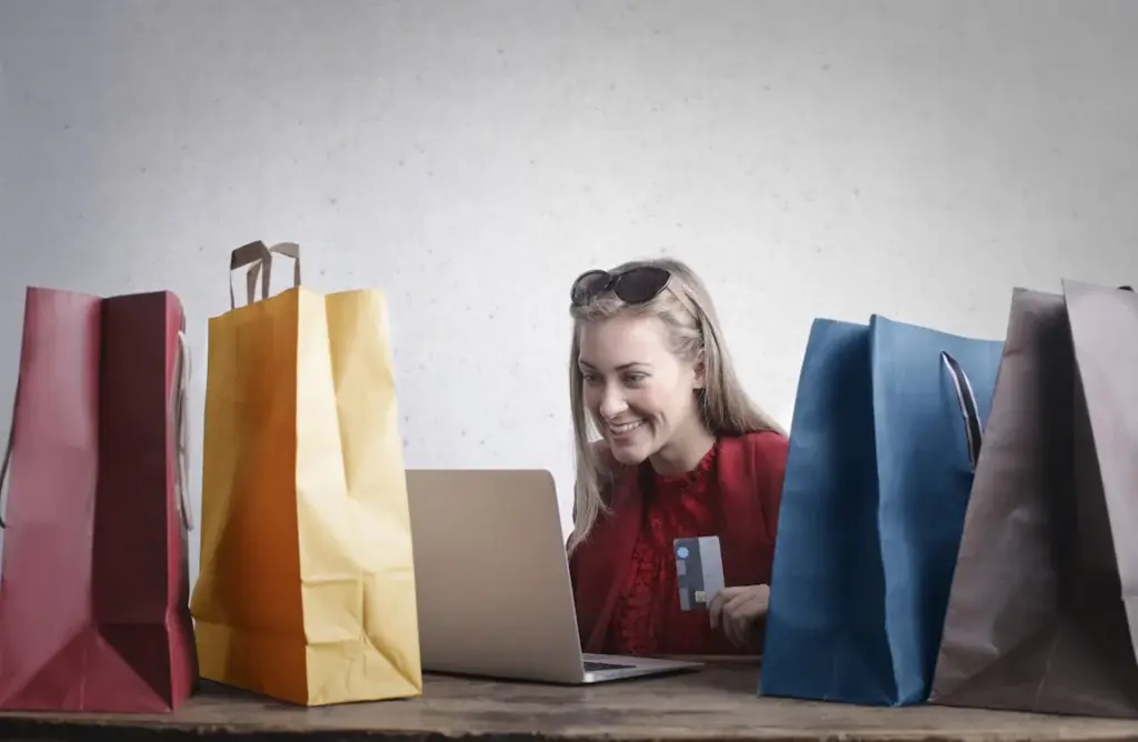 A person sits at a table with a laptop, surrounded by colorful shopping bags. They are holding a credit card and appear to be shopping online, showcasing how things replaced by technology have transformed traditional retail experiences.