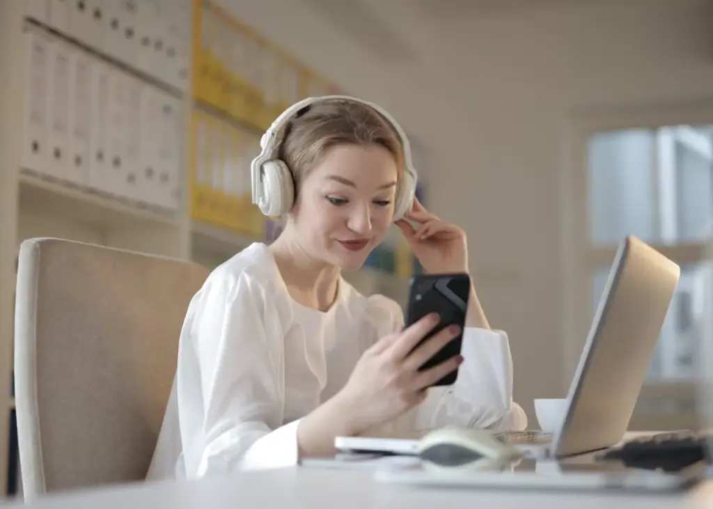 A woman wearing headphones sits at a desk with a laptop and files, looking at her phone and smiling, realizing that some things become less important as you get older.