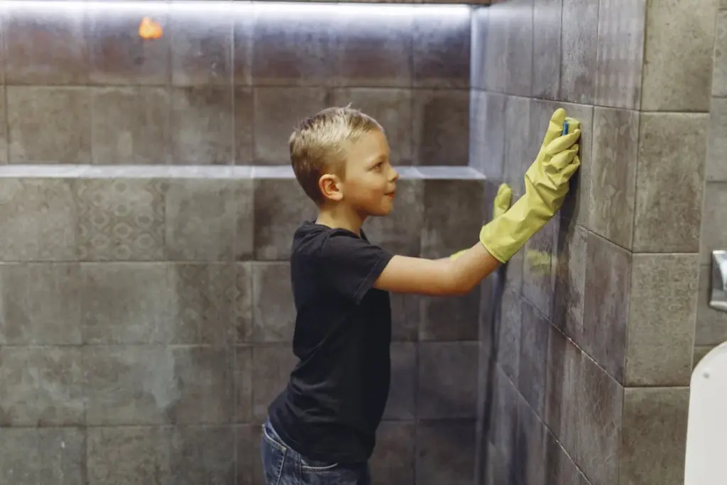 A child wearing yellow gloves cleans a tiled wall in a bathroom.