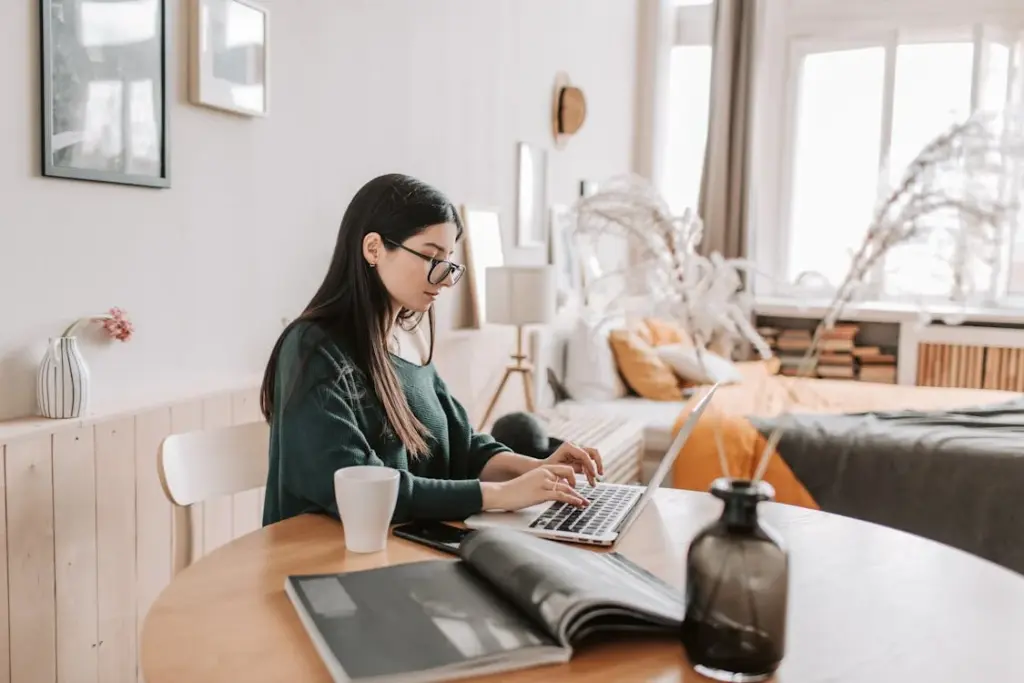 A person works on a laptop at a wooden table in a cozy bedroom, with a mug, notebook, and decorative items nearby.