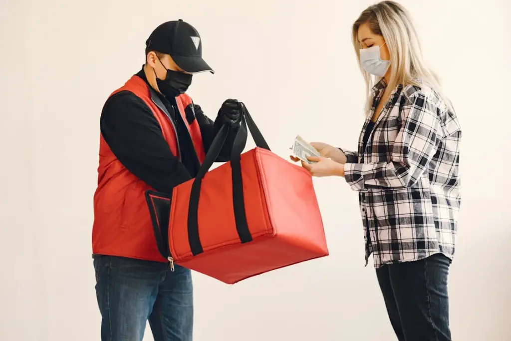 A delivery person hands a large red insulated bag to a woman while she gives him money. Both individuals are wearing masks.