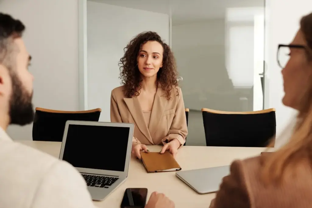 A woman sits at a conference table holding a notebook, facing two people with laptops open in front of them.