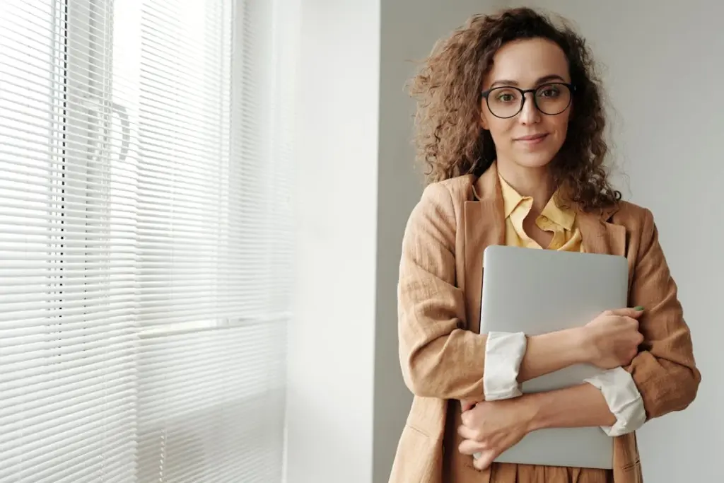 A person wearing glasses and a brown suit stands by a window, holding a closed laptop.