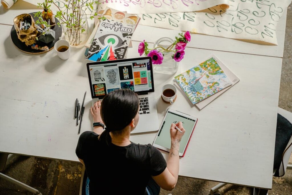 Person working on a tablet at a desk with a laptop, magazines, flowers, and coffee around them.