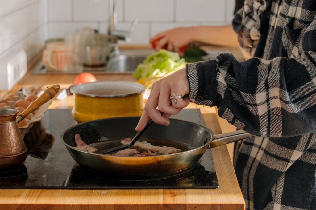 Person cooking bacon in a frying pan on a stovetop while another person preps vegetables in the background.