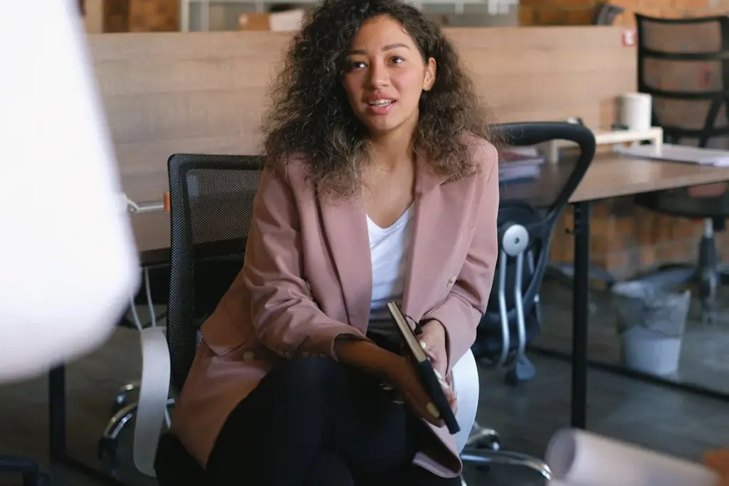 A woman with curly hair sits on an office chair holding a notebook, dressed in a pink blazer and white top in a modern office setting.