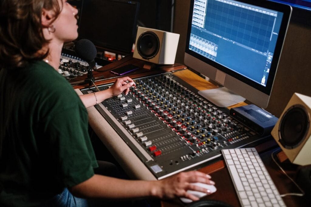 Person operating an audio mixing console and computer in a recording studio, surrounded by speakers and equipment.