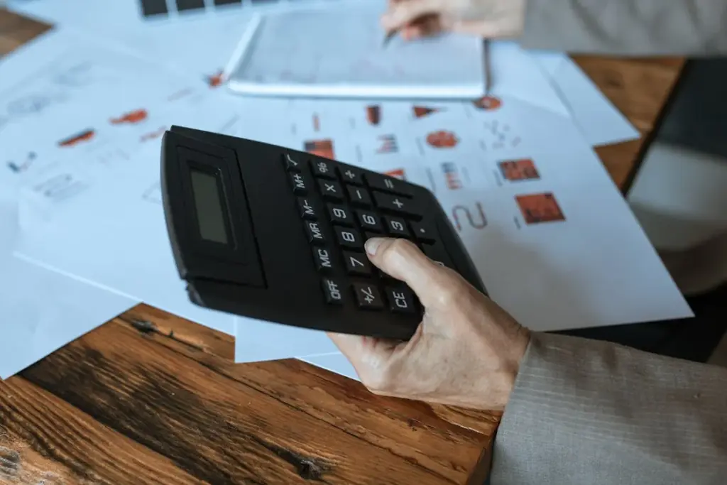 A person's hand holding a large calculator, with paperwork and a notebook on the wooden table in the background.