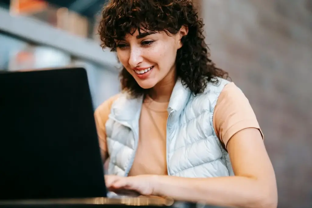 Person with curly hair, wearing a light puffer vest, smiles while using a laptop indoors.