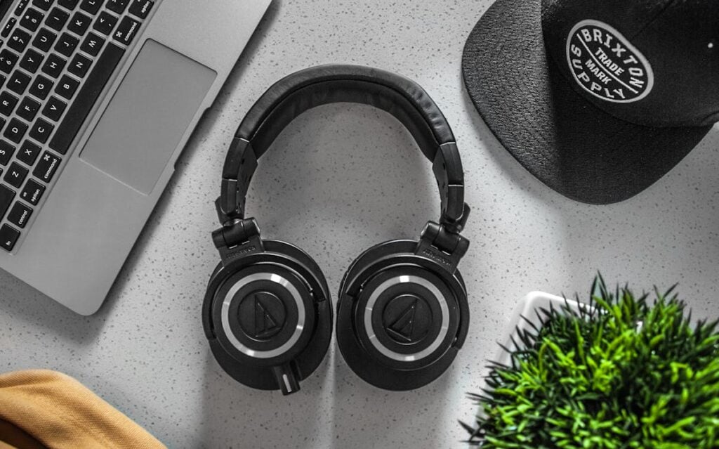 Overhead view of black over-ear headphones on a white desk, next to a laptop, a black baseball cap, and a small potted plant.