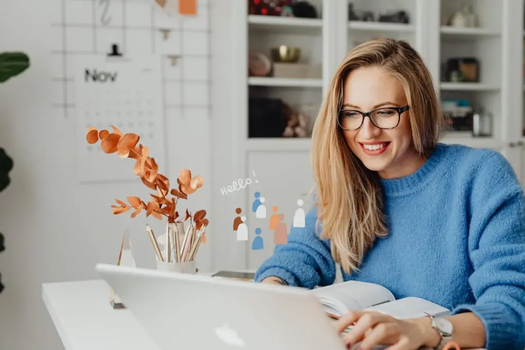 A woman in glasses and a blue sweater sits at a desk, smiling at her laptop. Behind her is a November wall calendar, and a vase with dried leaves is on the desk.