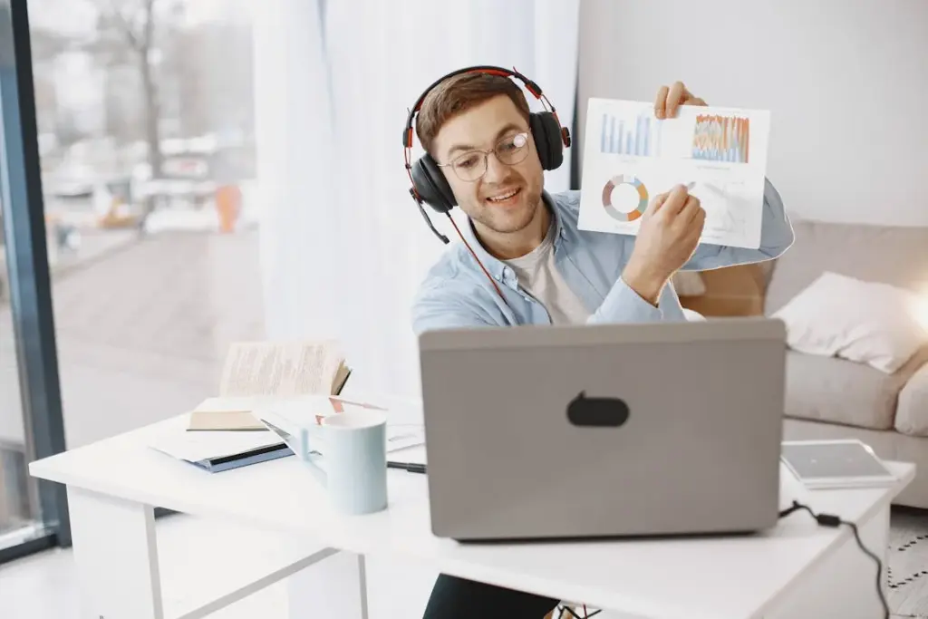 Person wearing headphones holds up charts during a video call in a bright room with a laptop and books on the desk.