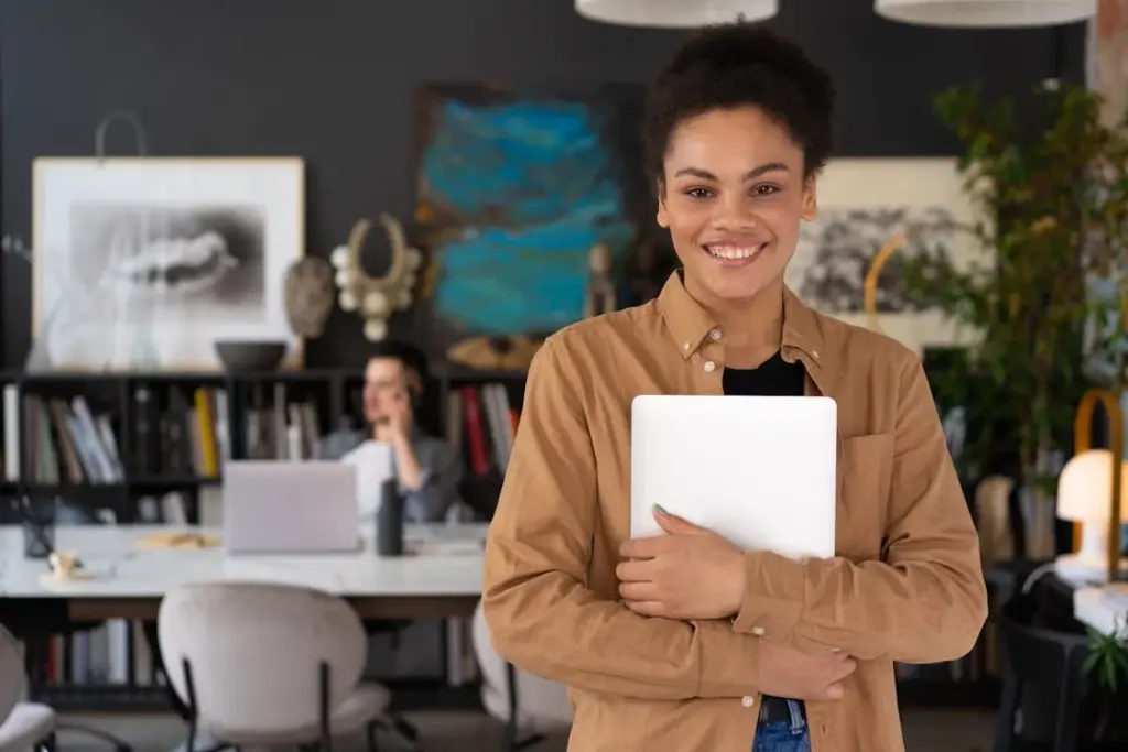 A smiling person holds a laptop in a modern office with bookshelves and art in the background. Another individual is working on a laptop at a desk behind them.