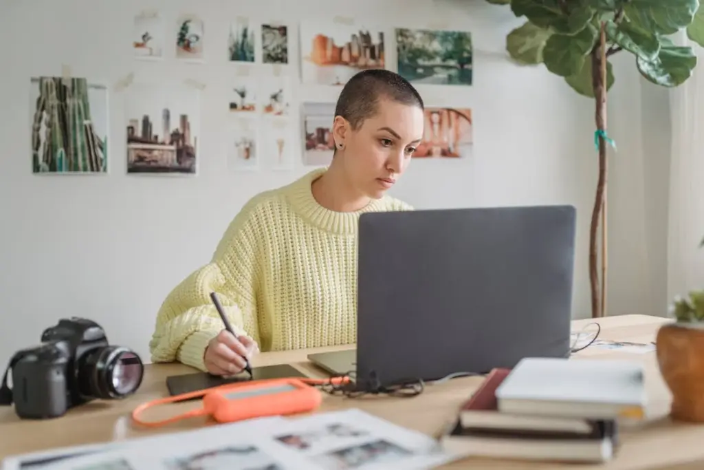 Person with short hair works at a laptop, surrounded by papers and a camera, in a home office decorated with photos and plants.