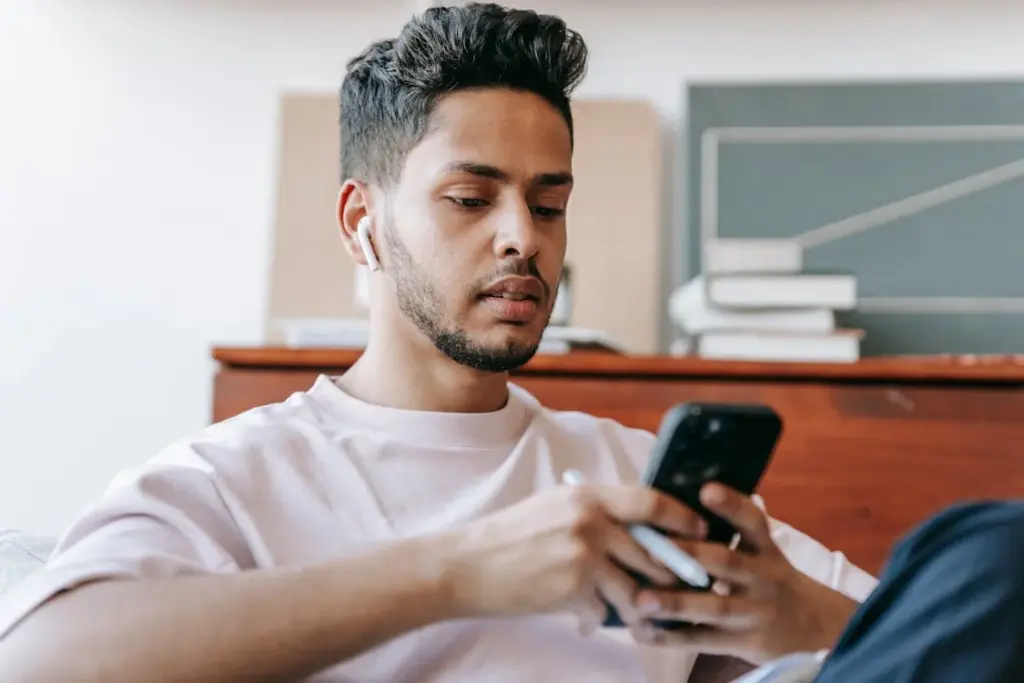 A person with short dark hair is sitting down, wearing a white t-shirt and wireless earbuds, and looking at a smartphone while holding a pen. There are books and a wooden cabinet in the background.