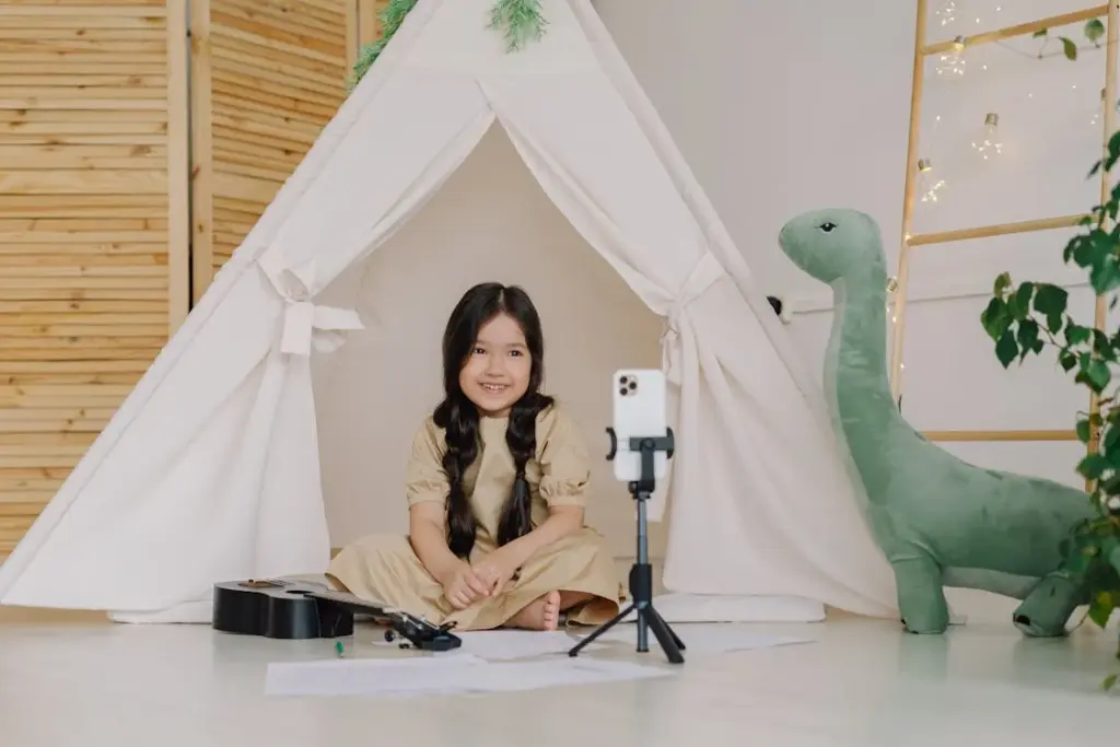 A girl sits inside a play tent with a guitar, papers, and a phone on a tripod. A stuffed dinosaur is beside her.