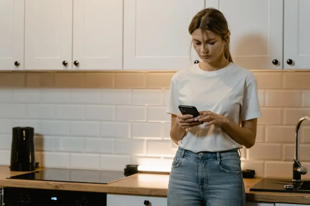 A woman in a white t-shirt and blue jeans stands in a kitchen, looking at her smartphone.