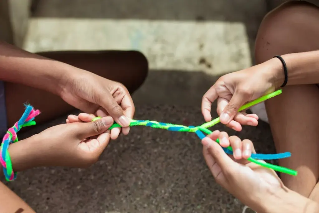 Four hands are twisting together colorful pipe cleaners, including green, blue, and pink, on a rough surface. One wrist has more pipe cleaners tied around it.