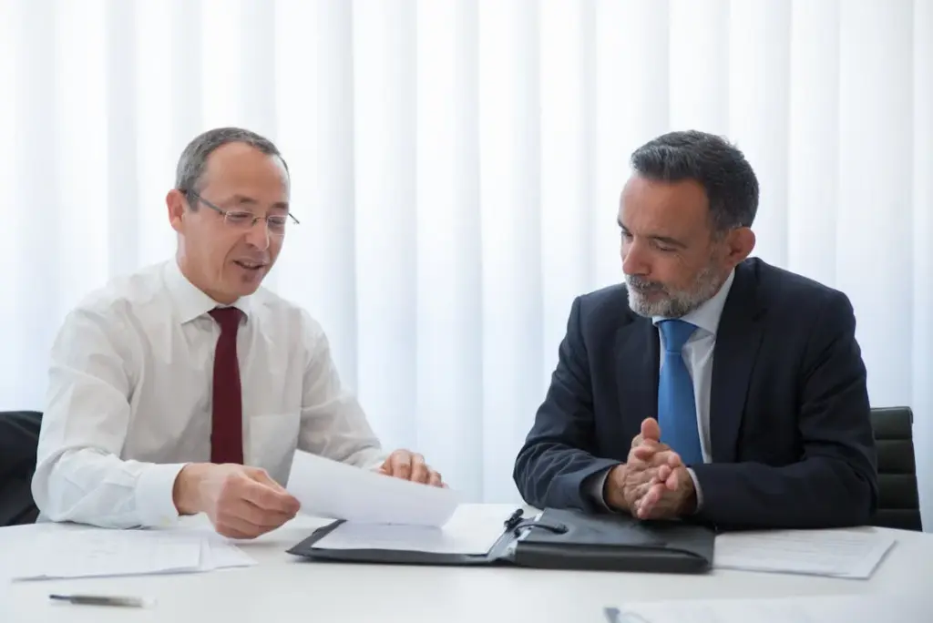 Two men in suits sit at a table with documents, engaged in discussion. One holds a paper while the other listens attentively, hands clasped.