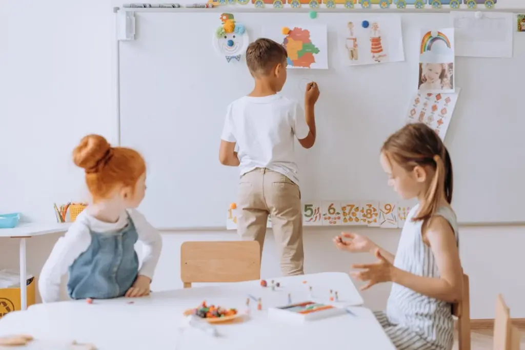 Three children in a classroom, one writing on a whiteboard and two sitting at a table with art supplies.