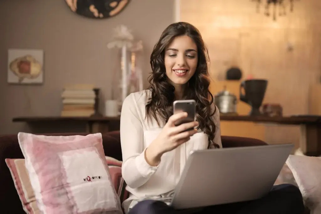 A woman sitting on a sofa is looking at her phone while holding a laptop on her lap in a cozy, neatly-decorated living room.