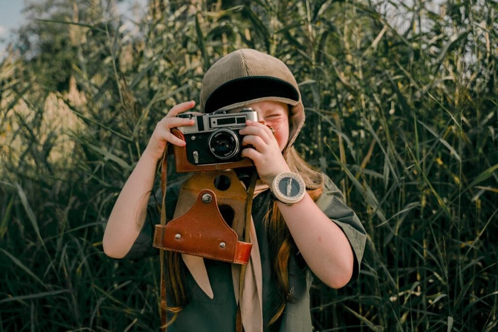 A person wearing a hat takes a photo with a vintage camera in a field of tall grass.